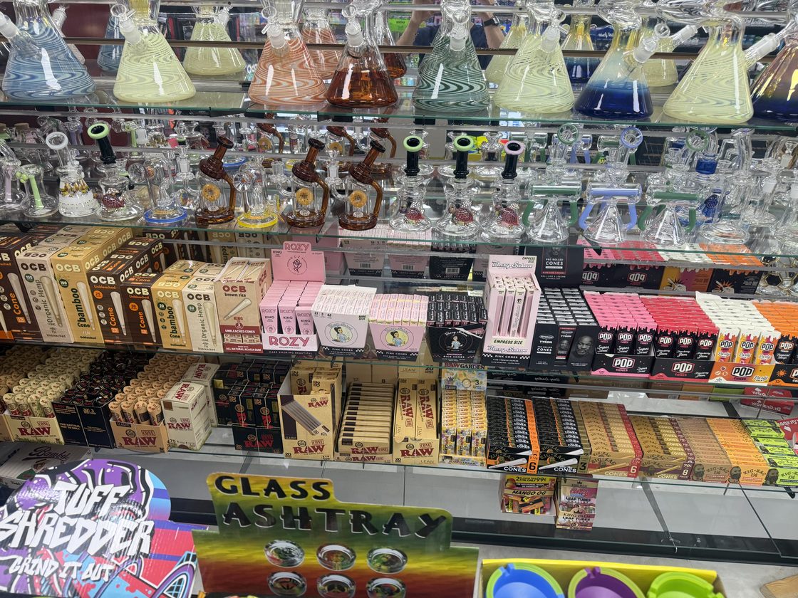 Inside the glass case: a row of beaker bongs on the top shelf, spoon pipes and recyclers below them, and a shelf of rolling papers and cones underneath.