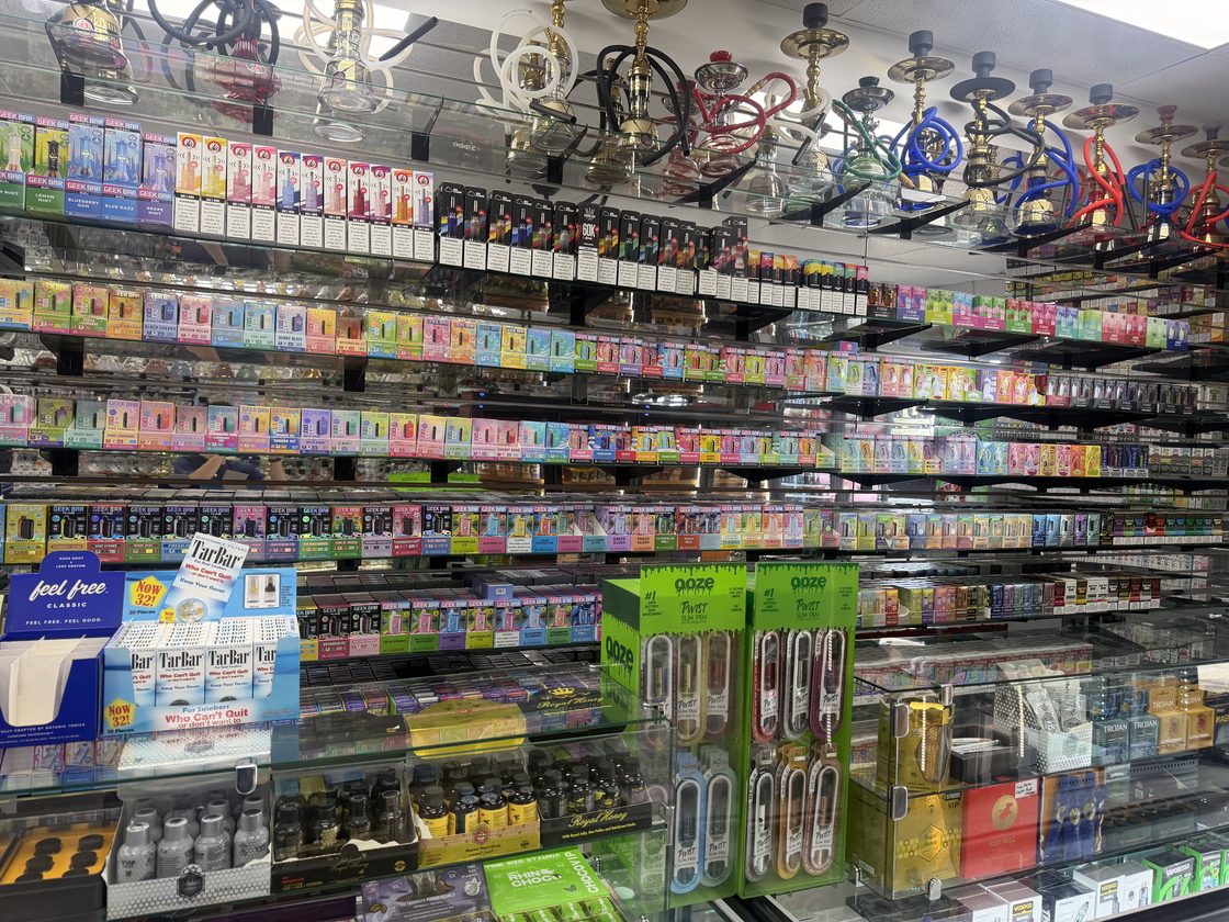The disposable vape wall: six shelves packed edge to edge with boxed disposables in every colour, e-liquid above them, and hookahs lined up along the top shelf.